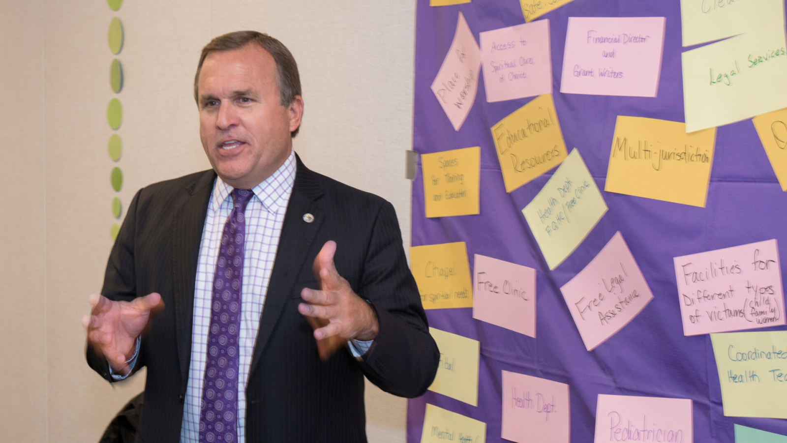Man in a suit speaking in front of a wall covered with colorful sticky notes featuring topics like victim support facilities.